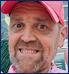 Close-up of a man with a beard, wearing a red baseball cap and pink shirt, smiling widely with an engaging, wide-eyed expression.