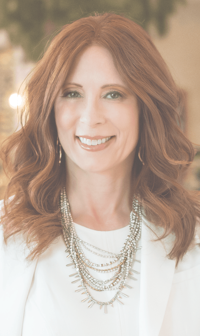 Smiling woman with wavy reddish-brown hair, wearing a white blazer and a multi-strand beaded necklace, looking directly at the camera.