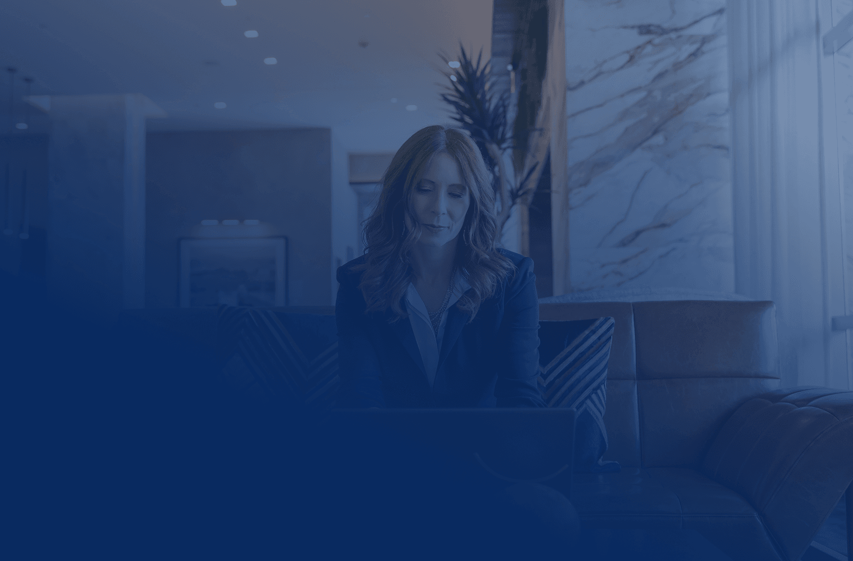 A professional woman with red hair intently uses a laptop while seated on a couch in a modern, blue-toned office lounge.